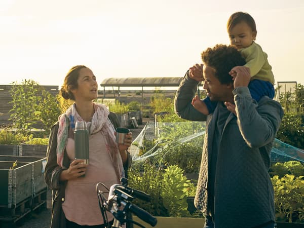 O familie de trei persoane într-o piață dintr-o grădină. Copilul stă pe umerii tatălui și mama ține în mână un termos.