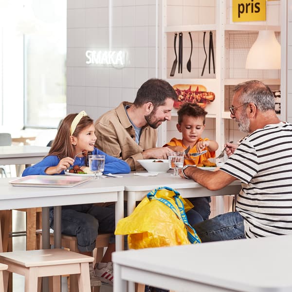 O familie formată din patru persoane stă la o masă într-un restaurant. Două copii și doi adulți mănâncă împreună. În fundal, se vede un semn luminos cu textul 