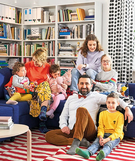 O familie mare - părinți și copii - adunată pe și în jurul unei canapele albastre; covor dungat și etajere pline în spate. 