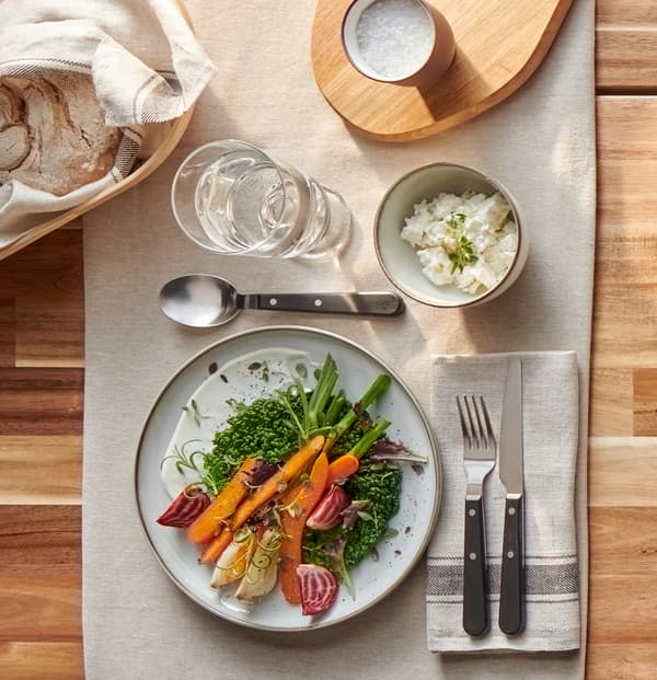 O farfurie rustică gri/maro, cu legume colorate, tacâmuri LIVNÄRA rezistente și un pahar cu apă pe o față de masă bej.