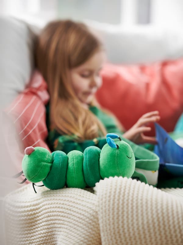 O fată care stă pe o canapea cu perne roșii, citind o carte lângă o jucărie muzicală sub formă de omidă de pluș verde numită KLAPPA.