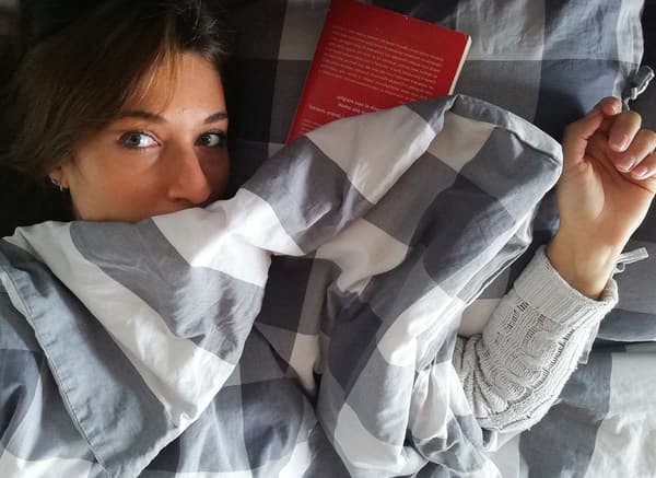 O femeie cu păr castaniu stă întinsă sub așternutul gri și alb. O carte roșie este așezată lângă capul ei.