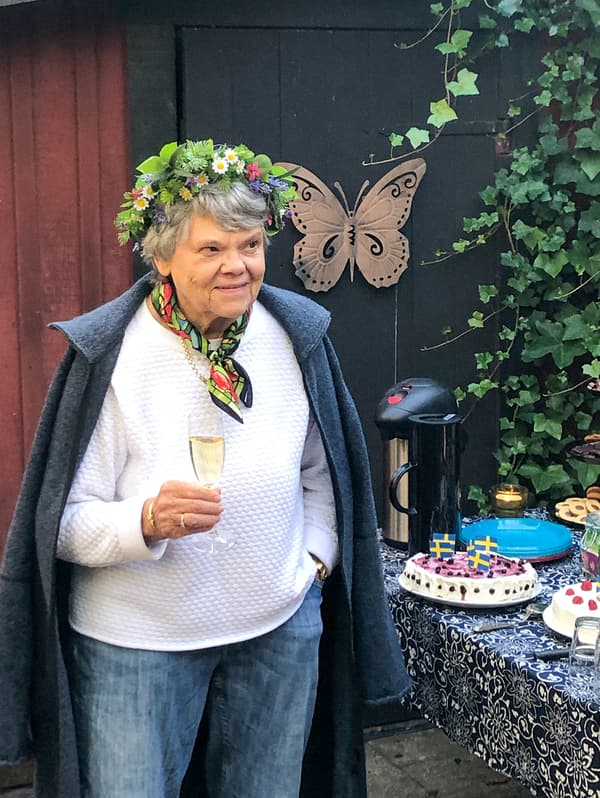 O femeie cu părul cărunt, cu o coroană de flori pe cap, stă în aer liber, ținând în mână un pahar de șampanie.