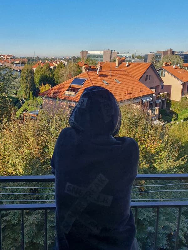 O femeie îmbrăcată cu un hanorac negru stă pe un balcon, privind la cartierul ei.