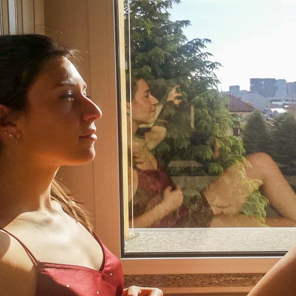 O femeie stă pe un pervaz însorit, cu ochii închiși. Reflecția ei este vizibilă în fereastra de lângă ea.