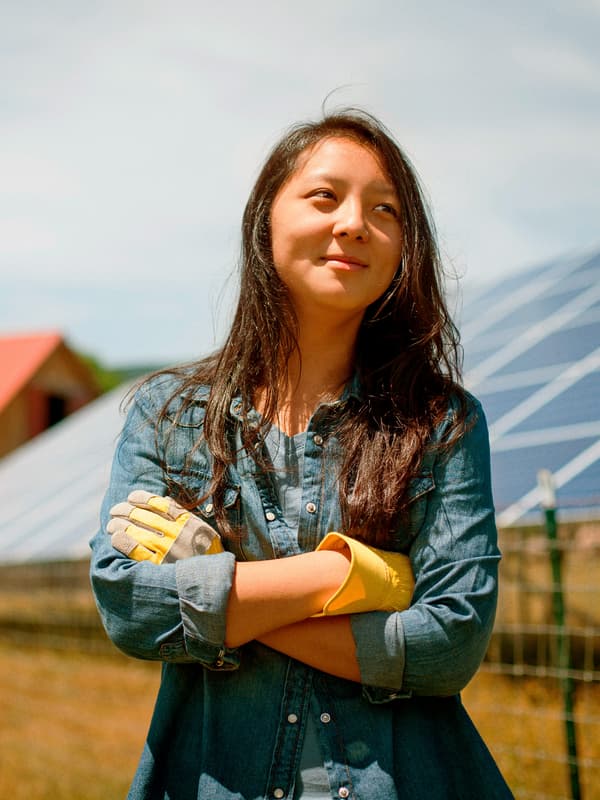 O fermieră zâmbitoare stă în fața a două case, una cu un acoperiș roșu, iar cealaltă cu un acoperiș cu panouri solare.