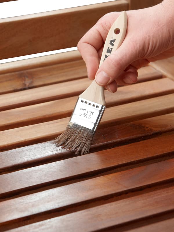 O mână care ține o pensulă acoperă șipcile din lemn ale unui obiect de mobilier de exterior cu baiț.