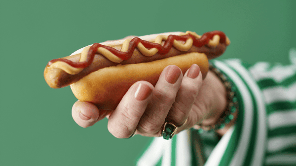O mână ține un hot dog cu muștar și ketchup, pe un fundal verde. Persoana poartă o brățară și un inel verde, iar mâneca hainei este în dungi albe și verzi.
