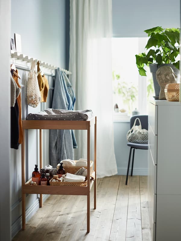 O masă de înfășat din lemn SNIGLAR cu o poliță de depozitare și un panou lung de cârlige din lemn albe pe peretele de deasupra sa.