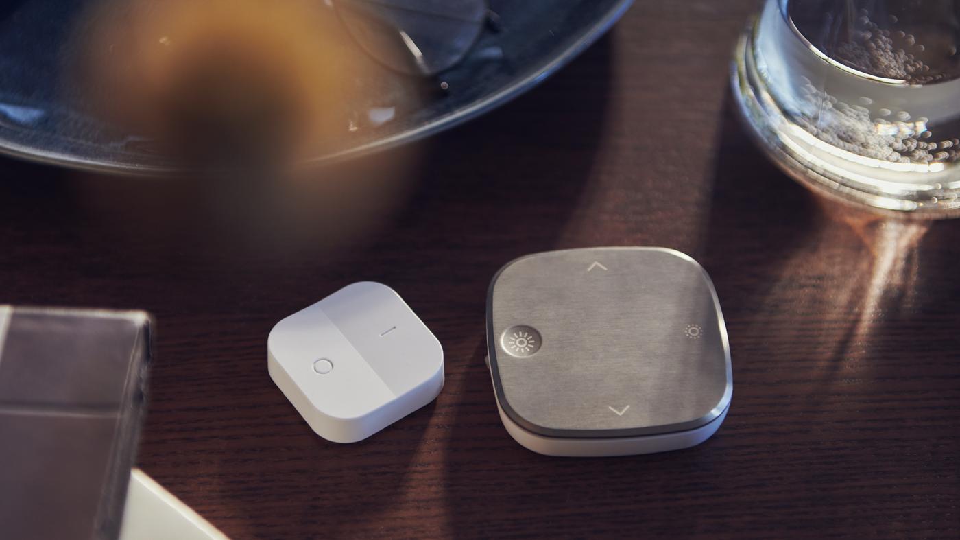 O masă maro pe care se află o farfurie rotundă, alături de un potențiator wireless TRÅDFRI alb și o telecomandă STYRBAR din oțel inoxidabil.