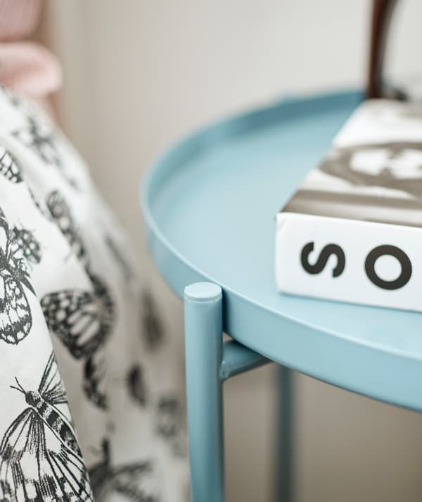 O masă rotundă din metal bleu cu o carte pe ea, lângă un pat acoperit cu o pilotă în alb și negru.