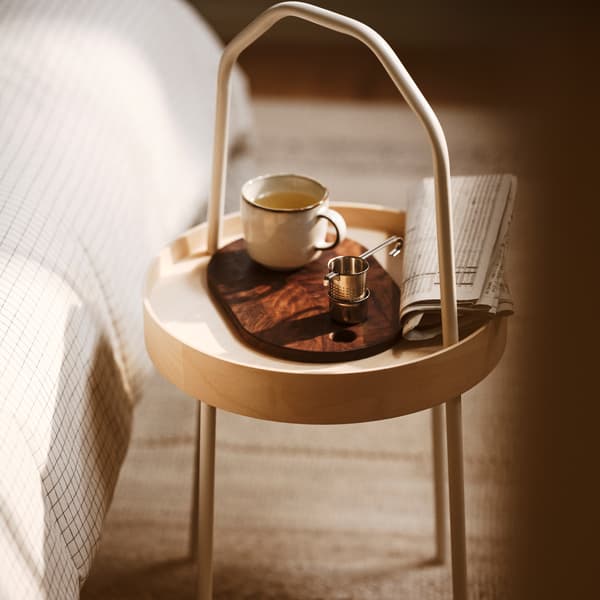 O măsuță de cafea BURVIK cu o cană GLADELIG gri așezată pe un tocător KRÖSAMOS. Măsuța de cafea este așezată pe un covor BRÖNDEN cu fir scurt.