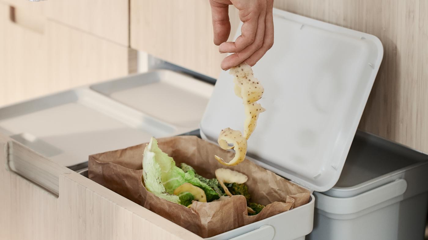 O persoană aruncă o coajă de legumă într-o pungă pentru deșeuri alimentare din hârtie maro într-un coș de sortare a deșeurilor HÅLLBAR aflat într-un sertar înalt MAXIMERA.