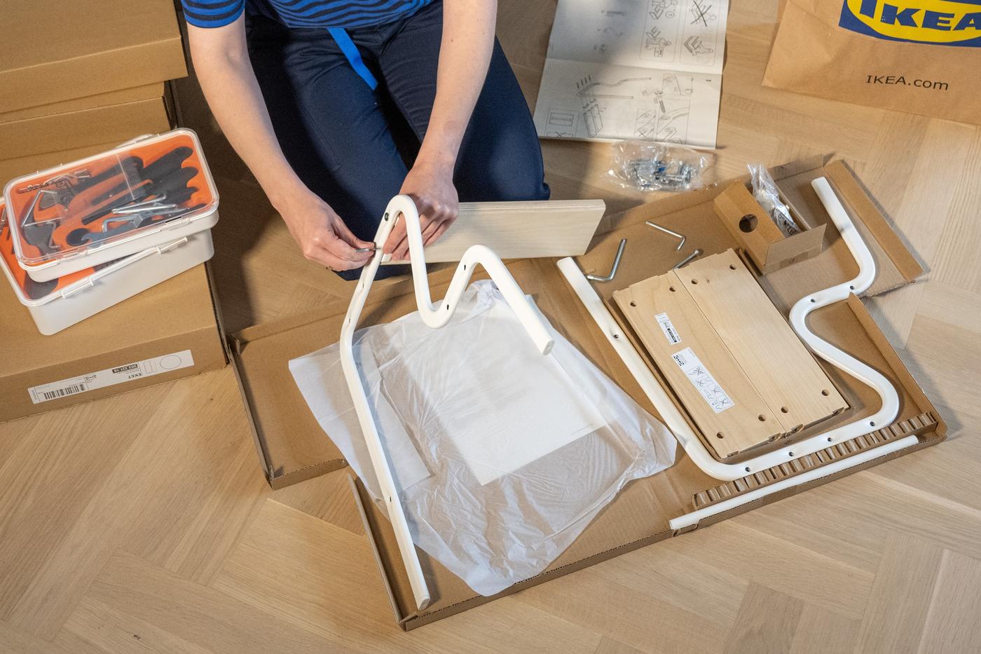 O persoană asamblează un obiect de mobilier IKEA, având în față piese din lemn și metal, șuruburi și o foaie cu instrucțiuni. Pe podea sunt cutii de carton și o cutie albă cu unelte.