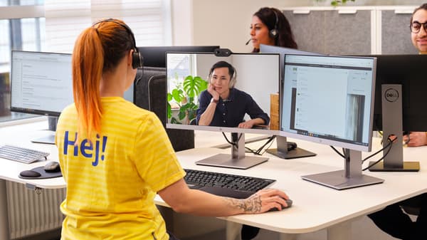 O persoană cu căști și tricou galben „Hej!” participă la o videoconferință pe două monitoare, așezată la birou. În fundal, două persoane lucrează la computere.