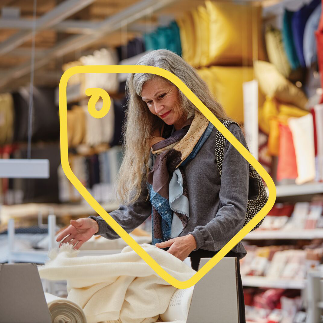 O persoană cu păr gri ține o bucată de material alb într-un magazin de textile, înconjurată de perne și produse textile expuse pe rafturi.