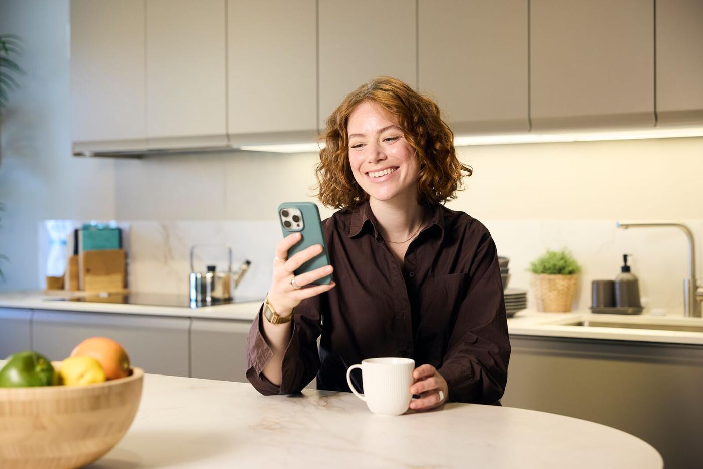 O persoană stă la o masă de bucătărie, ținând un telefon mobil în mâna dreaptă și o ceașcă albă în mâna stângă. În fundal se vede un blat de bucătărie cu diverse obiecte și un bol cu fructe pe masă.