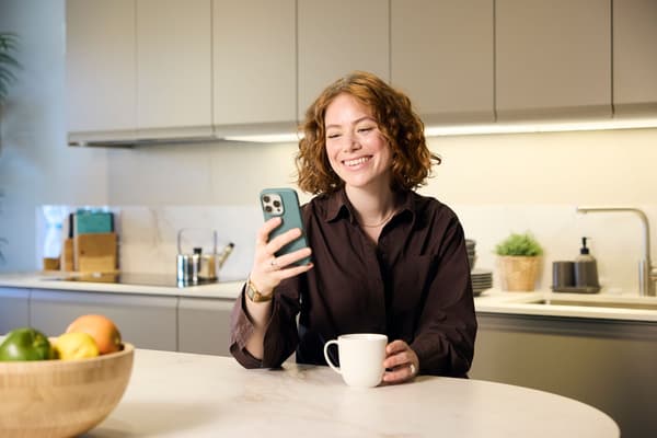 O persoană stă la o masă de bucătărie, ținând un telefon mobil în mâna dreaptă și o ceașcă albă în mâna stângă. În fundal se vede un blat de bucătărie cu diverse obiecte și un bol cu fructe pe masă.