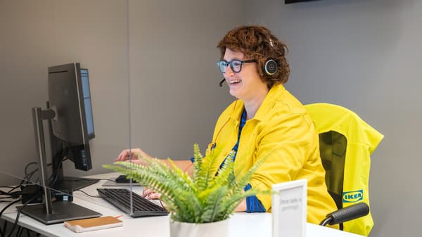 O persoană stă la un birou, purtând o cască cu microfon și o jachetă galbenă cu logo-ul IKEA. Persoana lucrează la un computer desktop. În prim-plan, pe birou, se află o plantă verde într-un ghiveci și un semn mic alb.