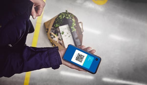 O persoană ține un telefon mobil în mână, pe ecranul căruia este afișat un cod QR. În fundal, pe podea, se află o pungă de hârtie cu plante și alte obiecte.