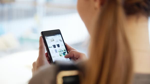 O persoană ține un telefon mobil în mână și navighează pe internet. Ecranul telefonului afișează imagini mici și text. Persoana este fotografiată din spate, iar părul său este prins într-o coadă de cal. Fundalul este neclar.
