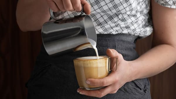 O persoană toarnă lapte spumat dintr-un recipient metalic într-o ceașcă de cafea, ținută în mână. Persoana poartă o cămașă albă cu carouri negre și un șorț gri.