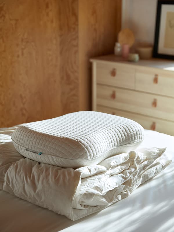 O pilotă și o pernă KLUBBSPORE sunt așezate una peste cealaltă pe o husă de saltea albă curată într-o cameră însorită.