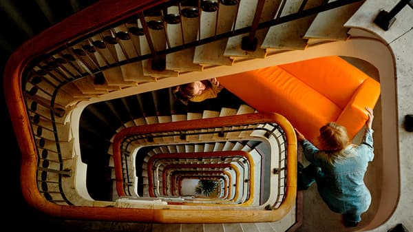 O scară în spirală într-o clădire cu multe etaje și un cuplu tânăr care transportă o canapea portocaliu aprins.