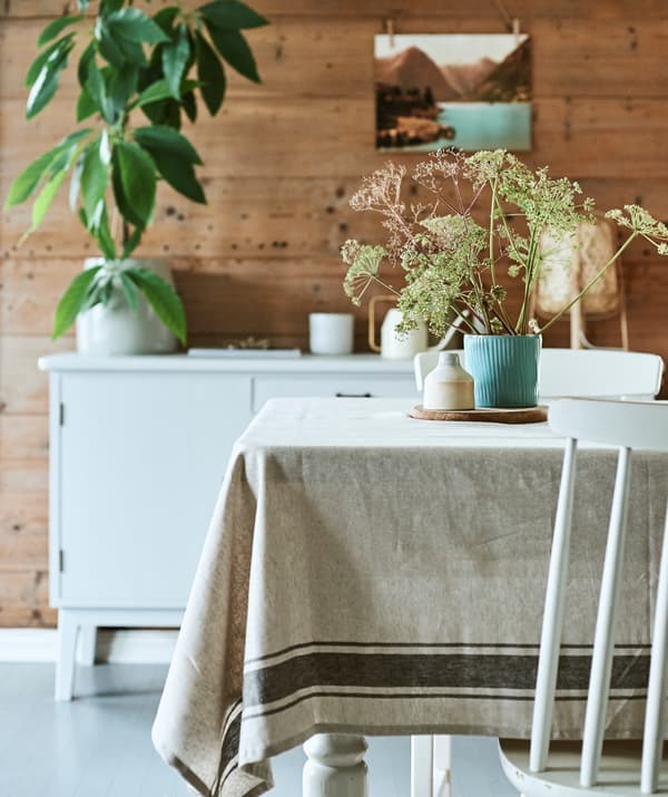 O sufragerie rustică, cu o masă din lemn acoperită cu o față de masă din in și un decor central din flori sălbatice într-un ghiveci albastru cu model.