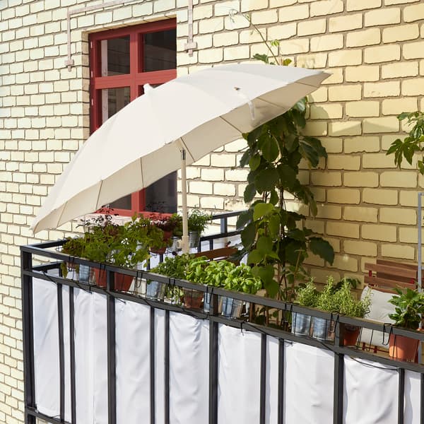 O umbrelă SAMSÖ/GRYTÖ, albă, înclinată pentru a bloca razele soarelui și a crea umbră, pe balcon. 