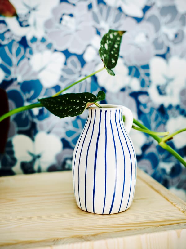O vază ceramică albă cu dungi neregulate albastre în care se află o plantă în fața unui tapet cu floral în combinația albastru și alb.
