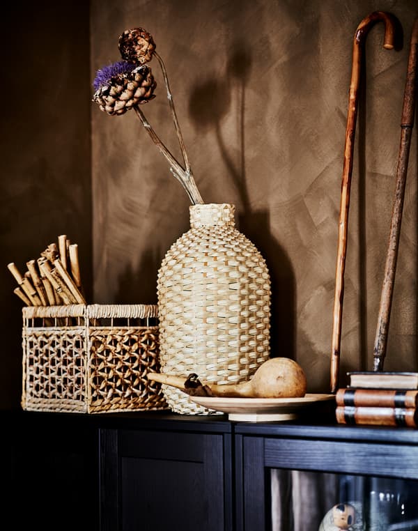 O vază decorativă KAFFEBÖNA realizată din detalii din bambus cu detalii deco din materiale organice se află pe partea superioară a unui corp HAVSTA maro închis.