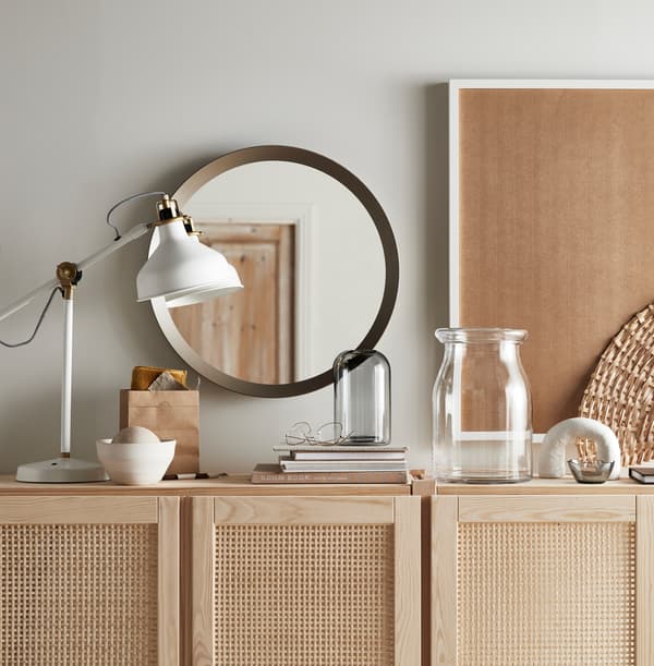 O veioză de birou RANARP albă, cărți, o oglindă rotundă și două vaze pe partea superioară a unui corp IVAR din lemn cu uși.