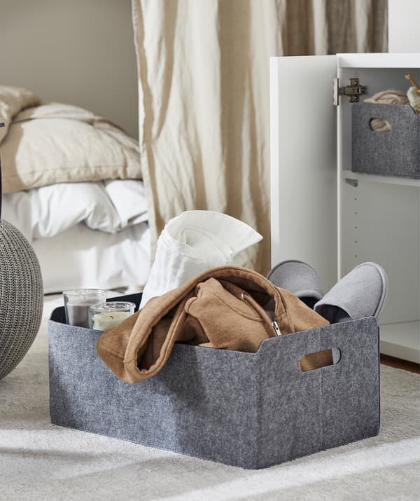 Obdĺžnikový plstený kôš položený na koberci v obývacej izbe, kôš s čajovými sviečkami, papuče, uterák a mikina.