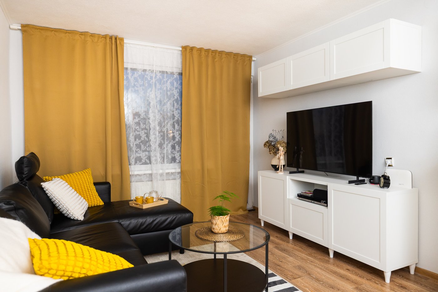Обновлённая гостиная с чёрным диваном, белой тумбой под ТВ и горчично-жёлтыми шторами. Пространство выглядит аккуратным, светлым и функционально организованным.
