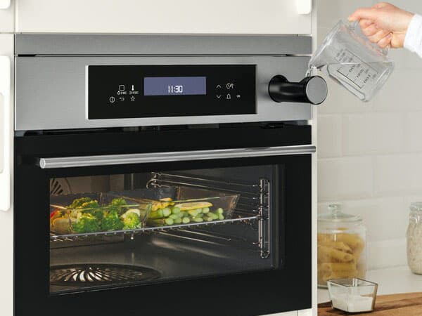oderne Küchenofen mit digitalem Display und Drehregler; eine Hand gießt Wasser aus einem Messbecher in den Ofen. Im Inneren garen Gemüse auf einem Blech. Im Hintergrund stehen Küchenutensilien wie Gläser und Behälter auf der Arbeitsplatte.