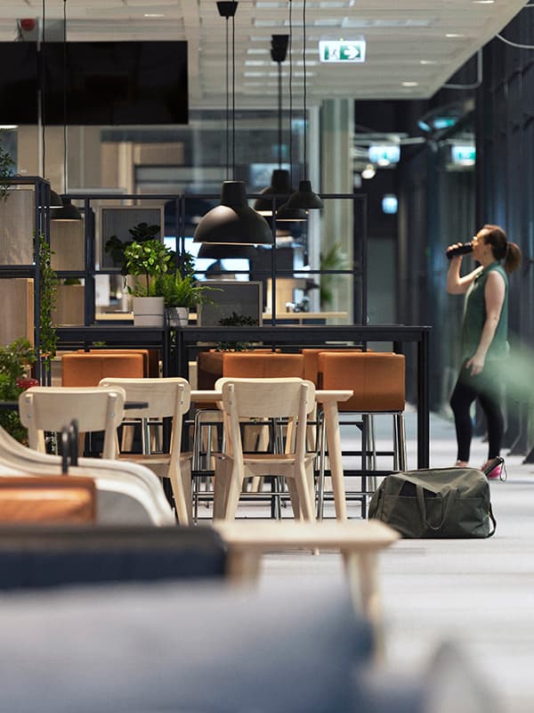 Öppet kontorsutrymme med bord, stolar, växter och hänglampor. En person står i bakgrunden med en väska på golvet.