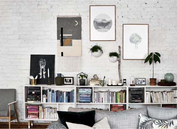 Œuvres d’art sur un mur de briques blanc, avec livres et plantes sur une étagère basse.