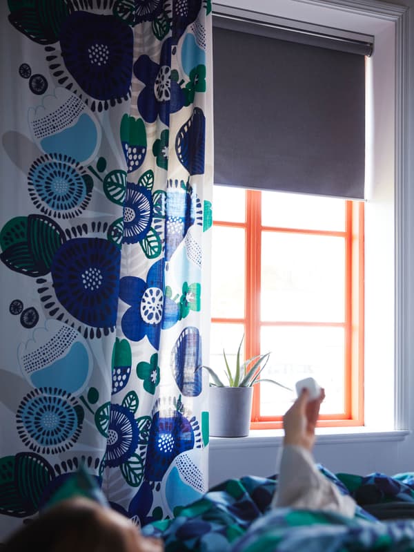 Okno so sivou zatemňovaciu roletou FYRTUR je čiastočne zakryté závesom s modro-zelenými kvetinami.
