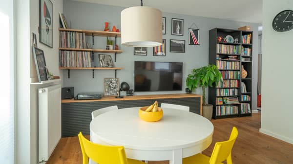 Okrągły, biały stół z zółtymi i szarymi krzesłami. Na drugim planie rozwiażanie z telewizorem na ścianie.