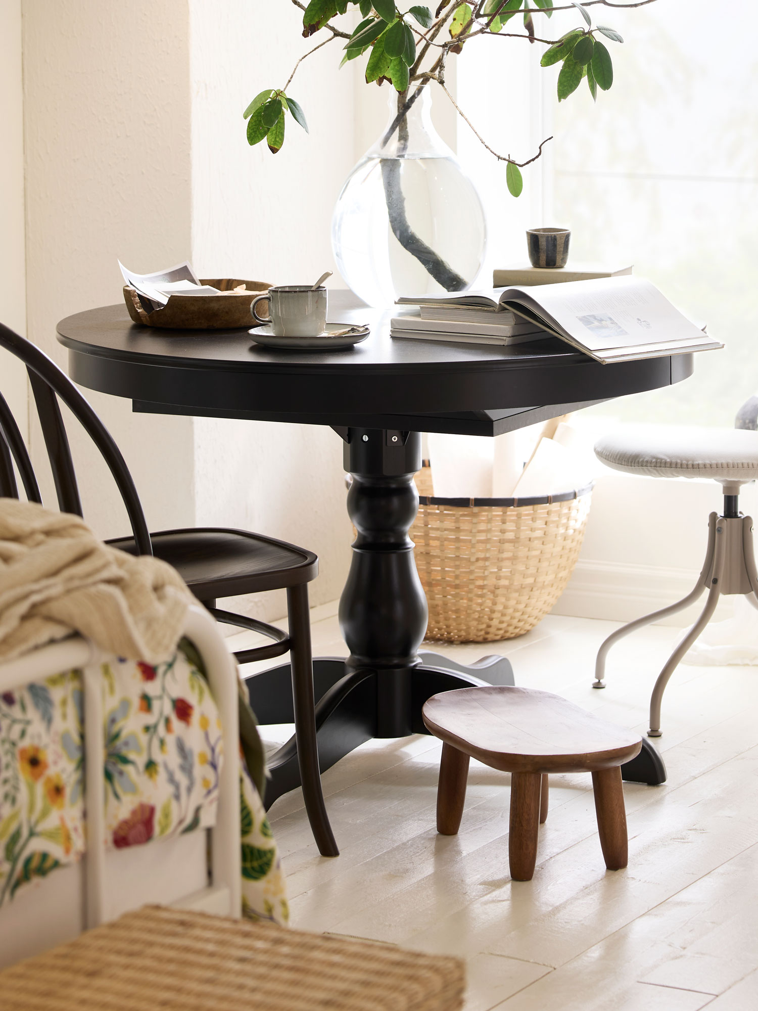 Okrugli crni stol s knjigom, šalicom na tanjuriću i drvenom zdjelom. Na stolu je velika staklena vaza s granom zelenih listova. Ispod stola je mala drvena klupica, a u pozadini košara i stolica.