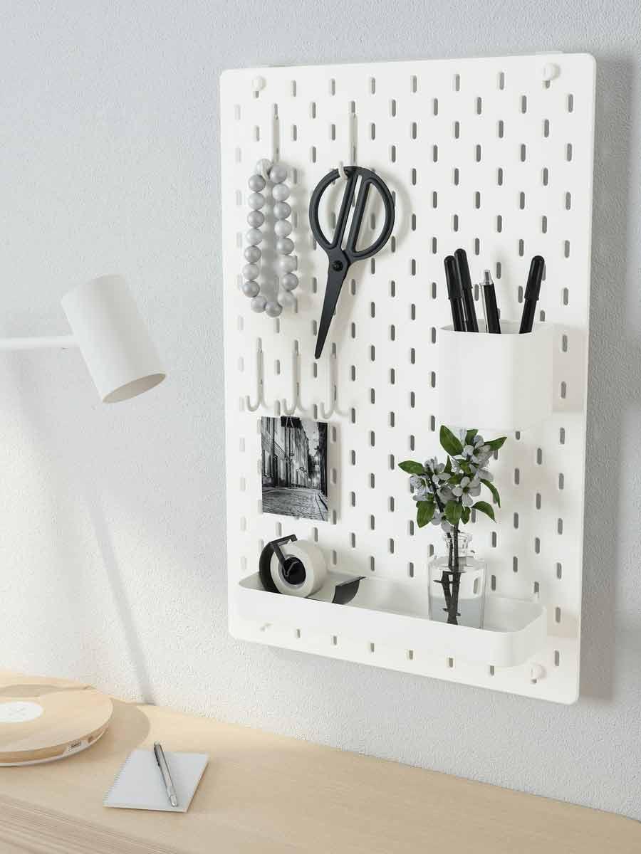 "On a white wall, there is a board hanging with items in it, such as scissors and pens.