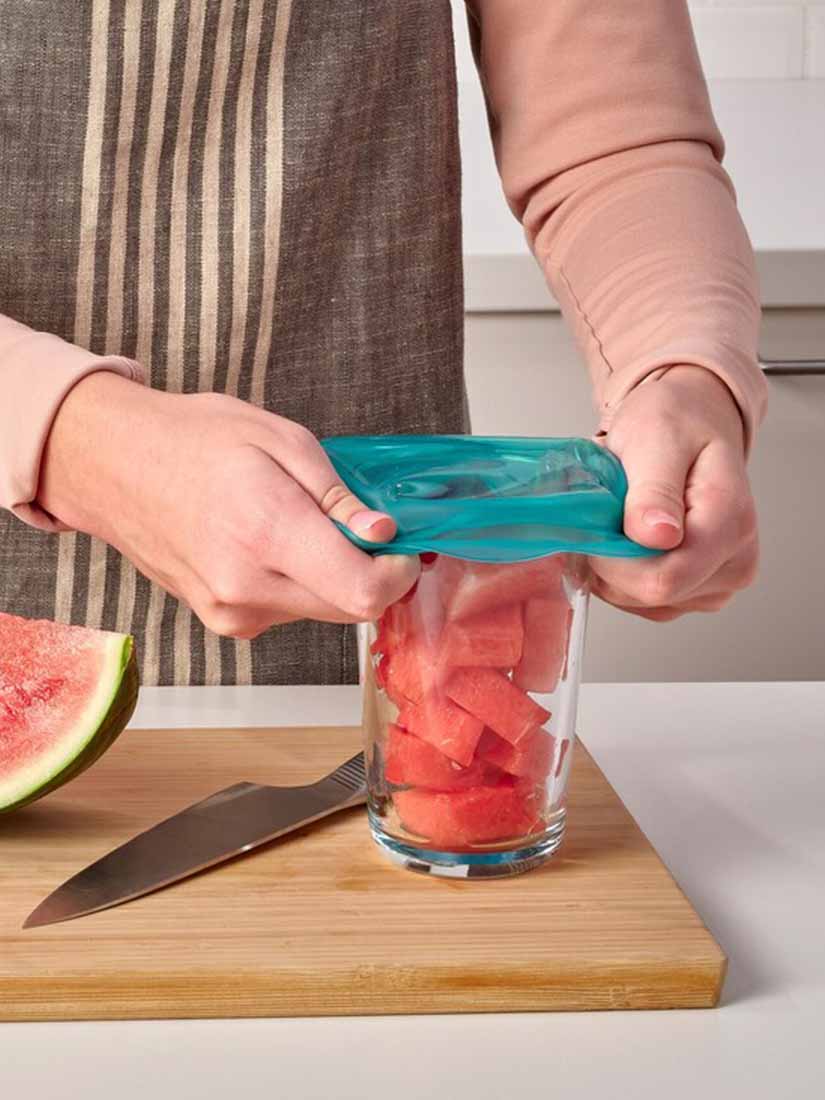 On a wooden chopping board, a person covers a glass of chopped watermelon with a reusable silicone ÖVERMÄTT food cover.