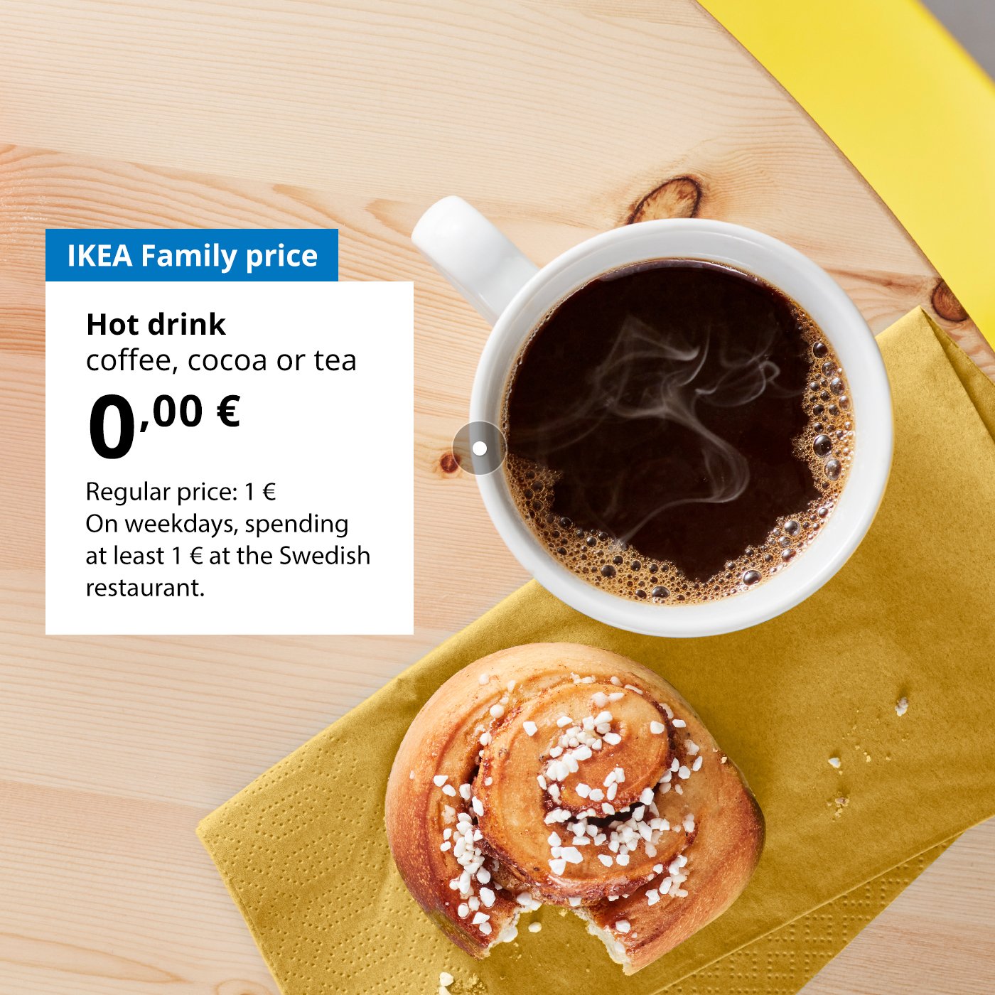 On a wooden table lies a half-eaten cinnamon bun and a mug of coffee. Next to the mug, against a white background, there’s an offer for IKEA Family club members: a hot drink (coffee, tea or cocoa) for 0 €, regular price 1 €.
