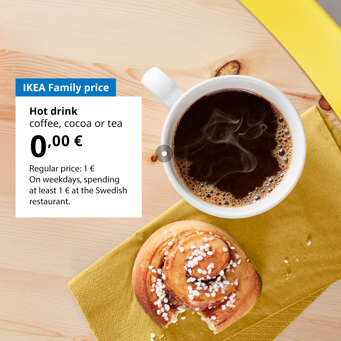 On a wooden table lies a half-eaten cinnamon bun and a mug of coffee. Next to the mug, against a white background, there’s an offer for IKEA Family club members: a hot drink (coffee, tea or cocoa) for 0 €, regular price 1 €.
