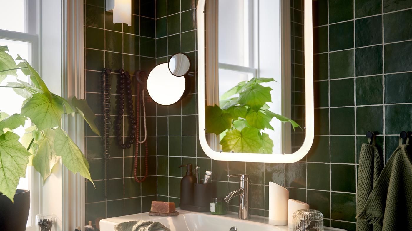 Onderdeel van een witte STORJORM spiegel met geïntegreerde verlichting boven een witte wastafel, plus een wandlamp met afneembare spiegel.