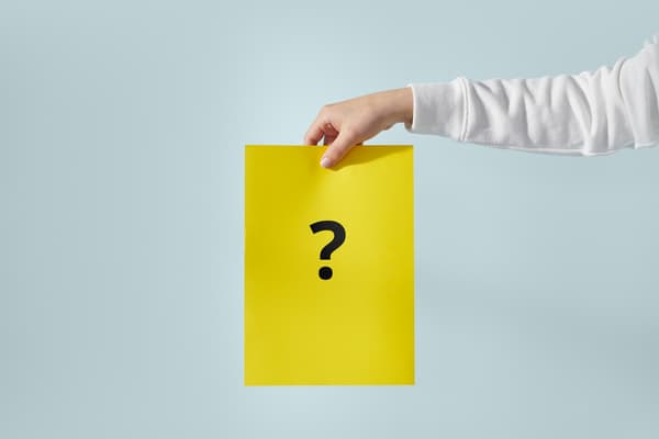 One hand holds a yellow A4 sheet with a question mark printed on it