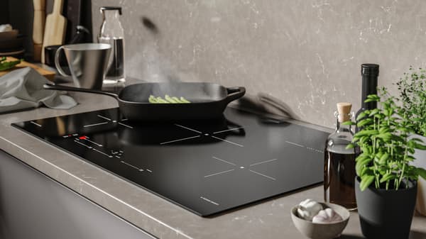 Op een zwarte SÄRKLASSIG inductiekookplaat staat een VARDAGEN grillpan waarop broccoli gaart. Achterin zie je keukengerei.