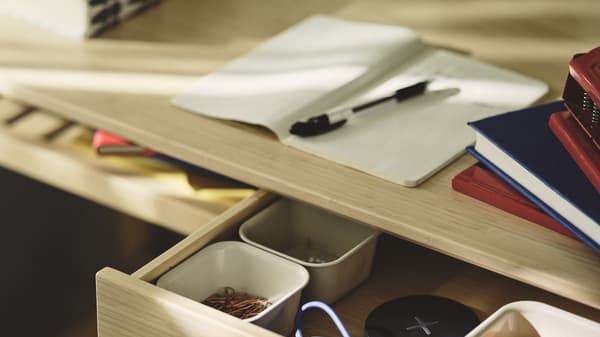 Open desk drawer with office supplies beneath a workspace featuring a notebook, pen, books, and stapler.