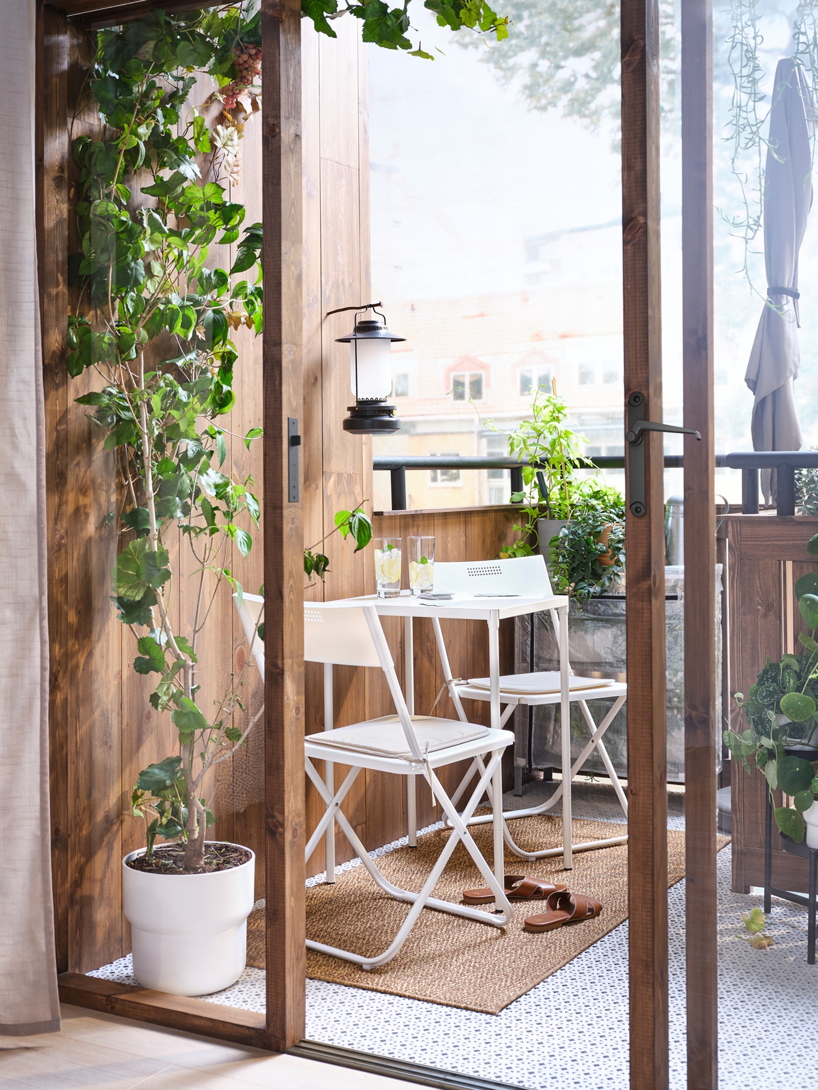 Open doors leading to a leafy balcony which has a white table and two chairs on it, with a lantern on the wall above.